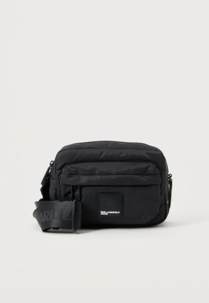 Petit sac bandoulière noir compact avec poche avant zippée et large bandoulière réglable sur fond blanc.