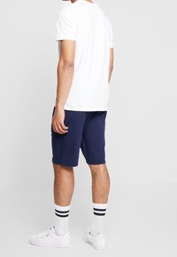 Homme portant un t-shirt blanc, un short bleu marine, des chaussettes blanches avec des rayures noires et des baskets blanches, se tenant contre un fond léger uni.