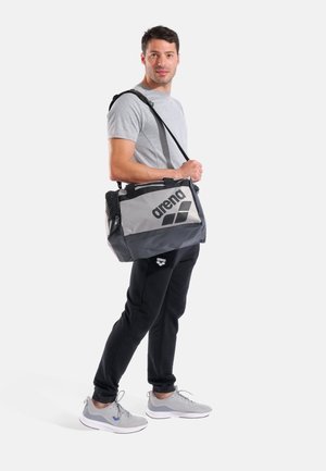 Mann in grauem T-Shirt und schwarzer Hose trägt eine graue und schwarze Arena-Sporttasche über der Schulter, steht vor weißem Hintergrund.