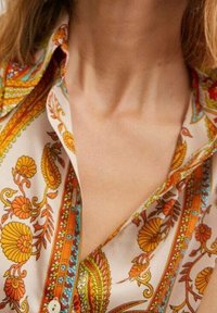 Femme portant une chemise à motifs floraux, déboutonnée, avec des designs orange, jaune et vert sur fond blanc, révélant son cou et le haut de sa poitrine.