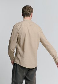 Beige langetermijnshirt met een gestructureerde kraag, knoopdetail op de rug en zijsplitten. De stof lijkt zacht met een lichte textuur.