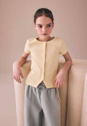 Joven chica con el pelo oscuro recogido, vestida con una camiseta amarilla de manga corta con botones y pantalones grises, apoyada en una superficie beige texturizada.