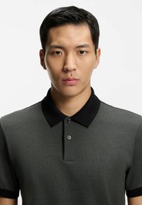 Tummanharmaa teksturoitu poolopaita, jossa on musta kaulus, kaksinappinen nappilista ja hillitty kohokuvio.