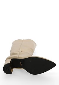 Beżowy skórzany kozak do kolan z czarną, spiczastą podeszwą i blokowym obcasem, przedstawiony od dołu na białym tle.