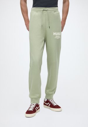 Uomo che indossa pantaloni della tuta Hugo verde chiaro con logo bianco e sneakers rosse con lacci bianchi su sfondo semplice.