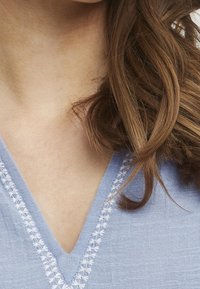 Vaaleansininen pellavapusero, jossa on V-aukko ja valkoinen kirjailu kaula-aukon reunassa; pehmeä tekstuuri ja rento istuvuus.