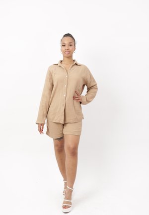 Mujer de pie con confianza, llevando una camisa beige abotonada y shorts a juego con sandalias blancas de tacón sobre fondo blanco.