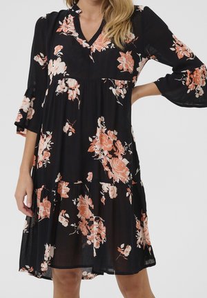 Frau trägt ein schwarzes knielanges Kleid mit pfirsichfarbenem und weißem Blumenmuster und 3/4 Glockenärmeln, steht mit einer Hand in der Hüfte.