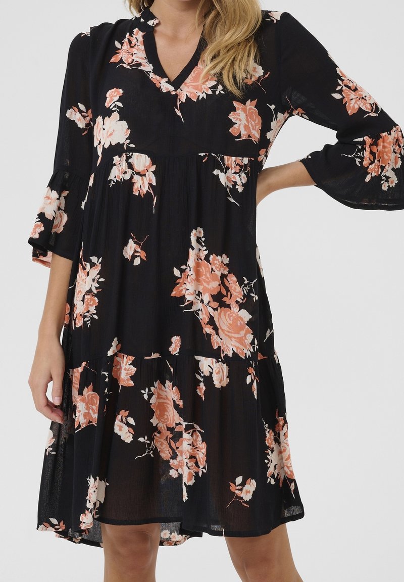 Femme portant une robe noire mi-longue aux genoux avec un imprimé floral pêche et blanc et des manches cloche 3/4, debout avec une main sur la hanche.
