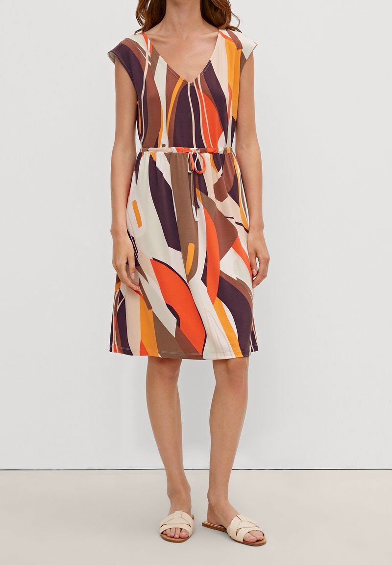 Femme portant une robe sans manches longueur genou avec un motif abstrait orange, marron et crème, taille marquée par un lien et sandales plates beiges.