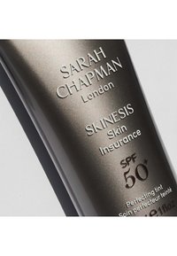 Graue Tube mit erhabenem Text; zeigt ‚SARAH CHAPMAN‘, ‚SKINESIS Skin Insurance‘ und ‚SPF 50+‘ auf einem metallischen Hintergrund.