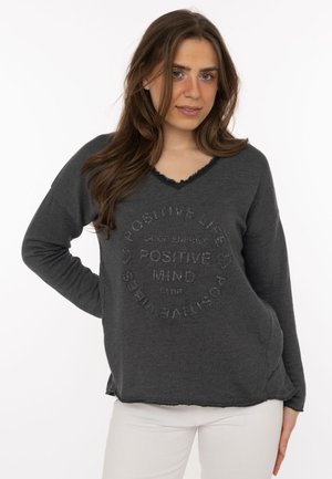 Young woman with long brown hair wearing a dark gray sweater with "Positive Mind" text and light-colored pants, standing with one hand on hip.