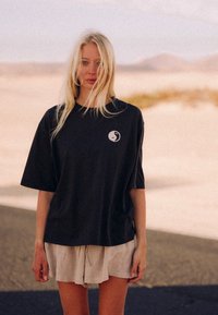 Czarna, oversize koszulka z krótkim rękawem i małym okrągłym logo na klatce piersiowej, w zestawie z jasnymi lnianymi szortami.