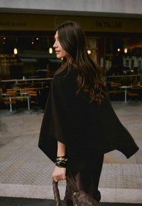Dark wool poncho drapes over shoulders, featuring a rounded hem. The individual wears a shiny black bangle and holds a braided brown handbag.