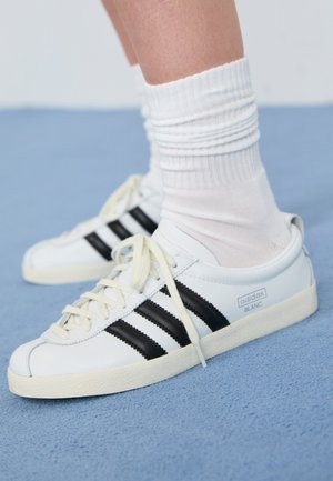 Fødder iført hvide Adidas Blanc sneakers med sorte striber og hvide crew sokker, stående på et blåt tæppe.