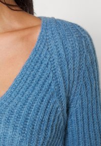 Vaaleansininen neulepaita, jossa on V-pääntie, ribatut pinnat ja pehmeä, pörröinen materiaali, jossa on voimakkaasti nostettuja ommelkuvioita.