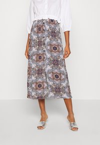 Femme portant un chemisier blanc court, une jupe midi à boutons à motifs, et des sandales métalliques ouvertes à enfiler, debout devant un fond uni.