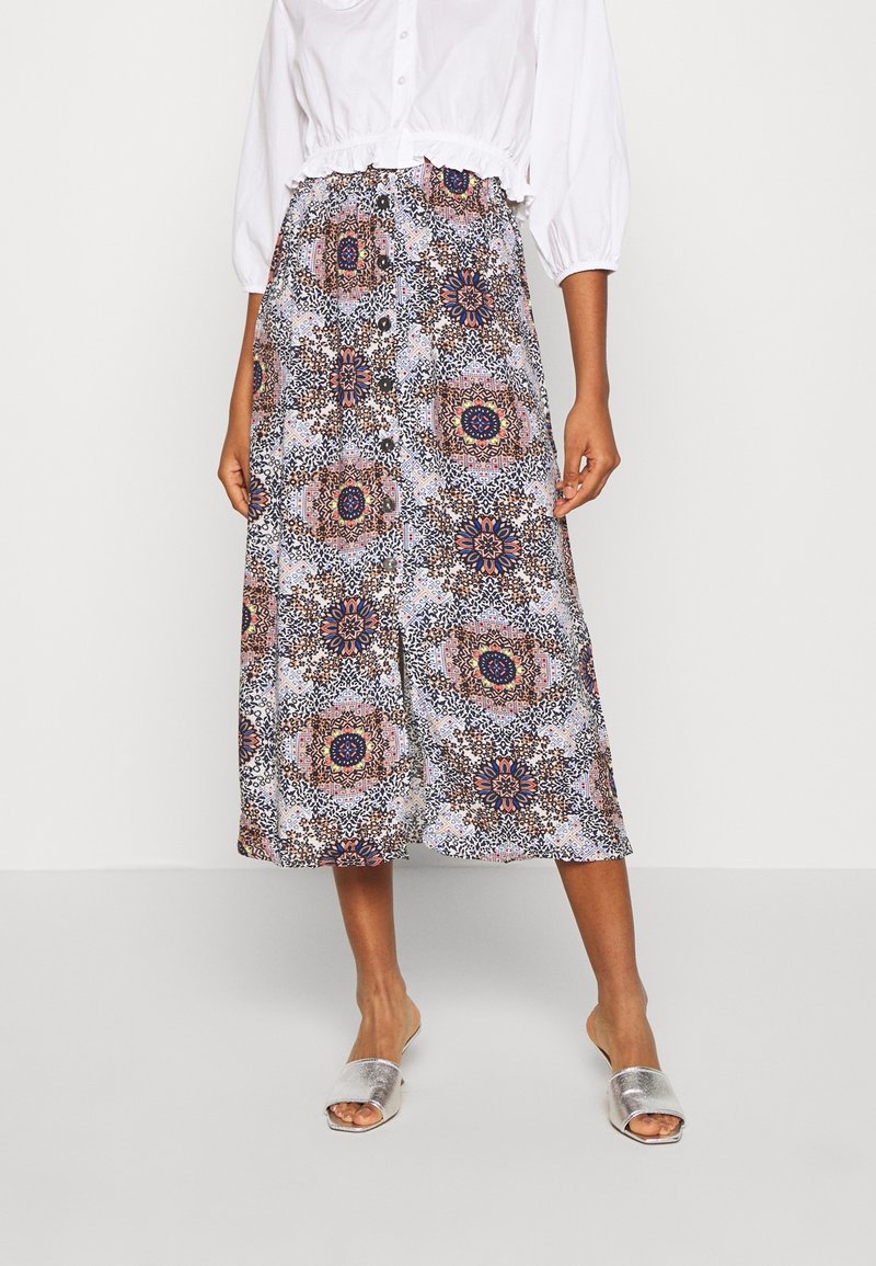 Femme portant un chemisier blanc court, une jupe midi à boutons à motifs, et des sandales métalliques ouvertes à enfiler, debout devant un fond uni.