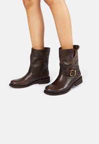 Bottes courtes en cuir marron avec un détail boucle, une finition texturée et un petit talon empilé. Fermeture éclair sur le côté pour un enfilage facile.