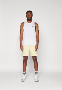 Λευκό αθλητικό μπλουζάκι με μαύρο λογότυπο Adidas, σε συνδυασμό με ανοιχτό κίτρινο σορτς. Γκρι αθλητικά παπούτσια με λευκές και μαύρες ριγωτές κάλτσες.