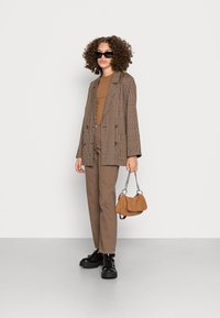 Blazer xadrez de dois botões em castanho, combinado com uma blusa castanha, calças bege e botas pretas de sola espessa. A segurar uma mala texturizada castanha.