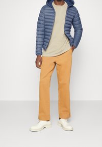 Navy puffy jacket with a hood, beige t-shirt, orange trousers, and white shoes. Jacket features quilted texture, trousers have straight fit.