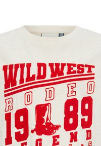 Koszulka w kolorze kremowym z czerwonym nadrukiem graficznym. Zawiera wyraźny tekst: "WILD WEST RODEO 1989 LEGENDS" oraz ilustrację kowbojskich butów.