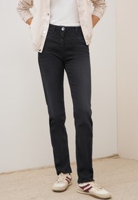 Zwarte rechtgesneden jeans met een standaard vijf-pocket ontwerp. Gedirigeerd met een lichtbeige jas en witte sneakers. Gladde denimtextuur.
