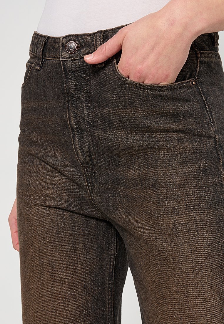 Hand in der vorderen Tasche einer dunkelbraunen Jeans mit sichtbaren Nähten und einem Metallknopfverschluss.