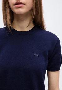 Νεαρό άτομο που φοράει ένα πλεκτό μπλουζάκι με κοντά μανίκια σε σκούρο μπλε χρώμα, με μικρό λογότυπο κροκόδειλου στο αριστερό στήθος, εμφανιζόμενο από τον λαιμό μέχρι το μέσο του κορμού.