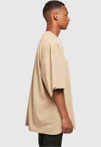 Beige suur T-särk, mis on valmistatud puuvillast, lühikeste varrukatega, ümarate kaelustega ja sirge hemiga. Ees teksti detail.