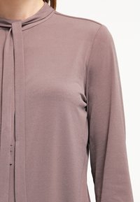 Langärmlige Bluse aus weichem Mauve-Stoff mit Kragen und gebundenem Halsdetail, glatte Textur und minimalistisches Design.