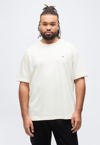 Άνδρας με κοτσίδες και γένια, που φοράει απλό κρεμ T-shirt και μαύρο παντελόνι, στέκεται μπροστά σε απλό λευκό φόντο.