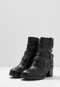 Black leather ankle boots with a rounded toe, layered straps, silver buckles, and a chunky heel. Features a zip closure and textured finish.