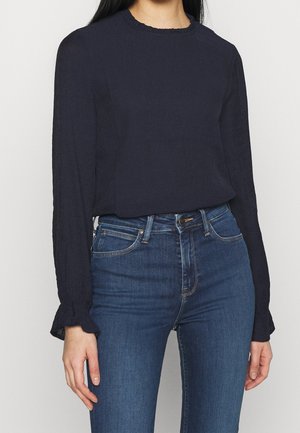 Frau trägt eine marineblaue Bluse mit langen Ärmeln und Rüschensäumen sowie hoch taillierte blaue Jeans, steht vor einem einfarbigen Hintergrund.