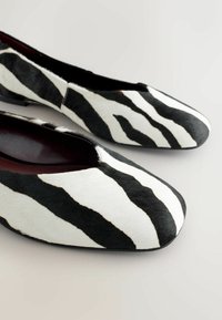 Paire de chaussures plates à rayures zèbre noires et blanches avec des bouts arrondis, présentées sur une surface claire.