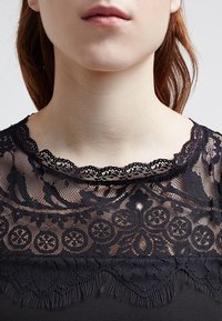 Czarna koronkowa bluzka z misternym kwiatowym wzorem, falbaniastym dekoltem oraz frędzlami na dolnym brzegu. Przezroczysta część górna i gęsta dolna.