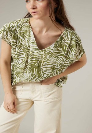 Femme portant un chemisier à manches courtes imprimé de feuilles vertes et blanches, avec un pantalon taille haute crème, posant avec une main dans la poche.