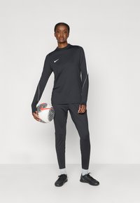 Μαύρο αθλητικό μπλουζάκι με μακριά μανίκια και εφαρμοστό παντελόνι με λευκές λεπτομέρειες, συνδυασμένο με μαύρες τάπες και κρατώντας μια κανονιστική ποδοσφαιρική μπάλα.