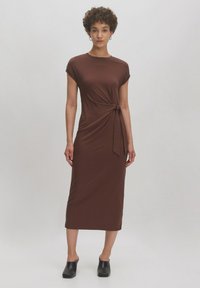 Braunes, knielanges Kleid aus glattem Stoff, mit kurzen Ärmelchen und einem gebundenen Detail an der Taille. Kombiniert mit schwarzen Slipschuhen.