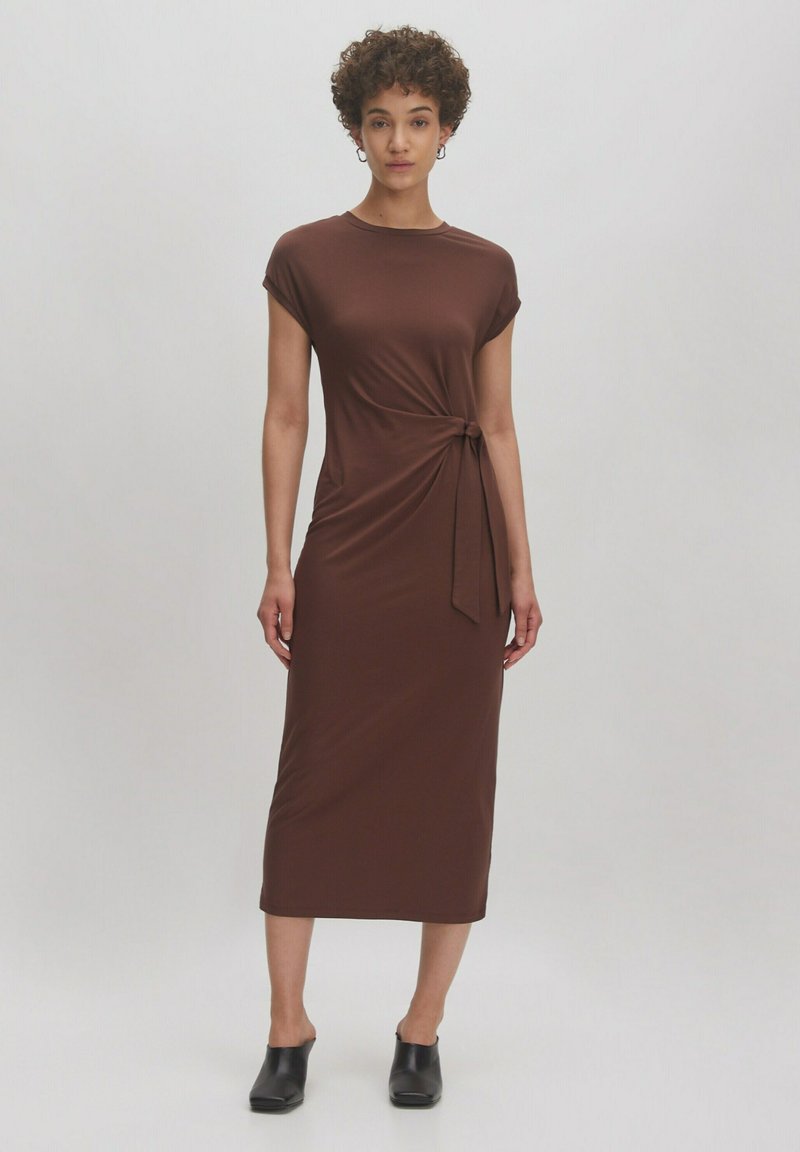 Braunes, knielanges Kleid aus glattem Stoff, mit kurzen Ärmelchen und einem gebundenen Detail an der Taille. Kombiniert mit schwarzen Slipschuhen.