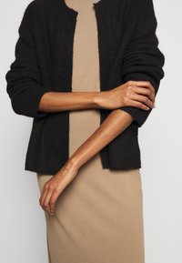 Cardigan nero lavorato a maglia con una vestibilità rilassata e una texture a costine, abbinato a un vestito attillato beige che mette in evidenza un tessuto liscio.