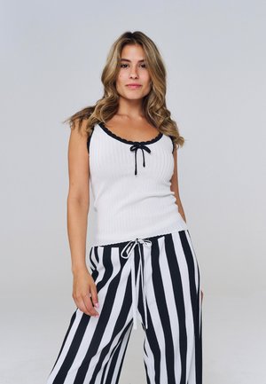 Jeune femme aux cheveux ondulés et lâchés, portant un haut blanc sans manches avec un bord noir et un pantalon à rayures verticales noir et blanc.