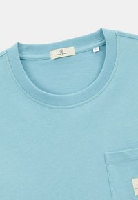 T-shirt en coton bleu clair avec un col rond, un col côte et une poche plaquée sur le devant. Étiquette visible à l'intérieur du col.
