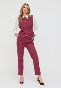 Burgunderrotes, maßgeschneidertes Weste- und Hosen-Set aus strukturiertem Stoff. Darunter ein weißes Hemd mit klassischem Kragen und goldenen Knöpfen. Schwarze Absatzschuhe.