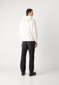 Felpa bianca con cappuccio dal tessuto liscio, abbinata a pantaloni neri dalla vestibilità comoda. Il modello indossa calzature nere, vista da dietro.