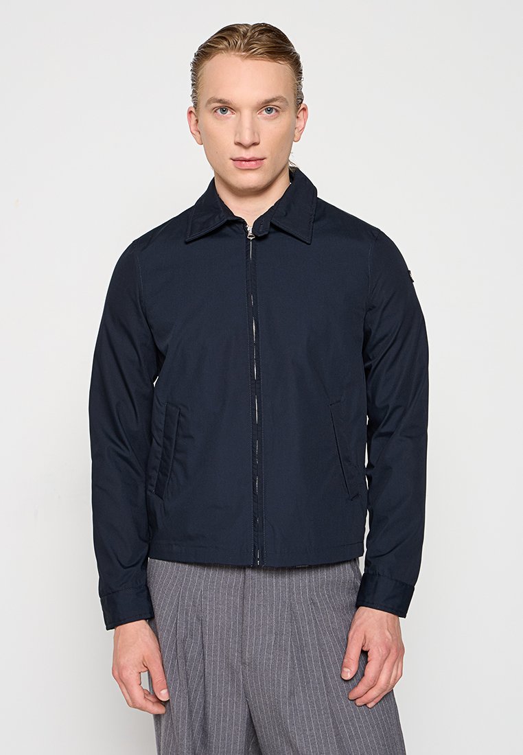 Veste zippée bleu marine avec un col, des poches avant et une texture lisse. Portée sur un pantalon gris à rayures, le modèle se tient contre un fond neutre.