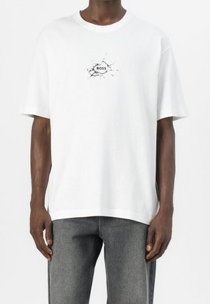 Mężczyzna w białym oversize T-shircie z wzorem pękniętego szkła i napisem "BOSS" na piersi, w połączeniu z szarymi jeansami.