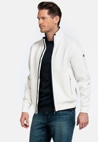 Witte zip-up jas met zwarte accenten, voorzien van een opstaande kraag, zijzakken en geribbelde manchetten, gedragen over een zwart shirt en blauwe jeans.