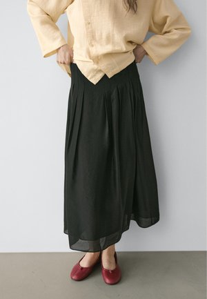 Person trägt ein beigefarbenes Hemd mit Knöpfen, einen schwarzen plissierten Midirock und rote Ballettschuhe, steht vor einer schlichten hellen Wand.